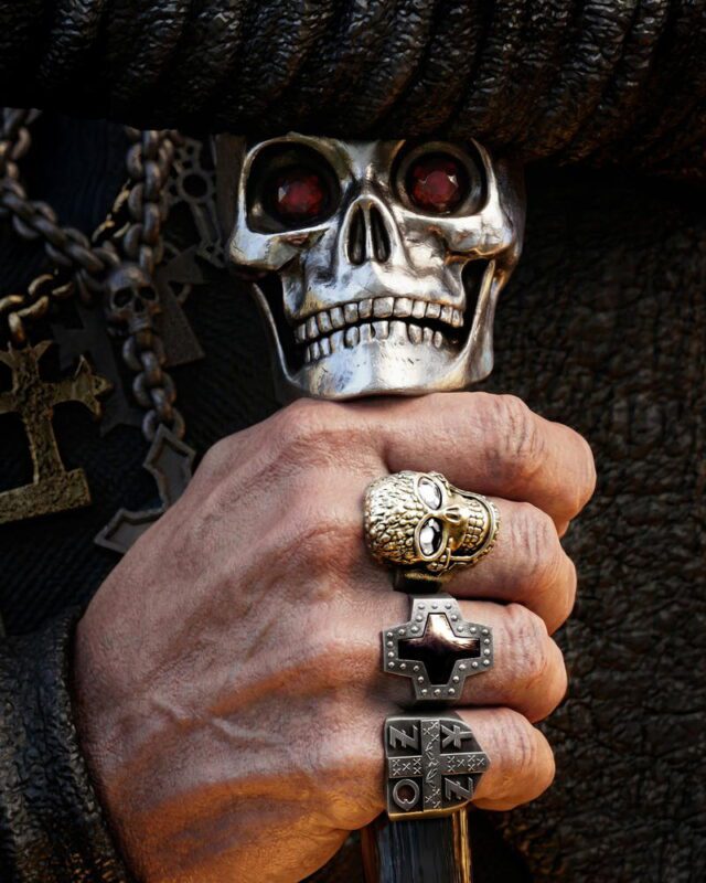 Close-up of a hand gripping a silver skull ring with red gemstone eyes, surrounded by gothic jewelry and chains.